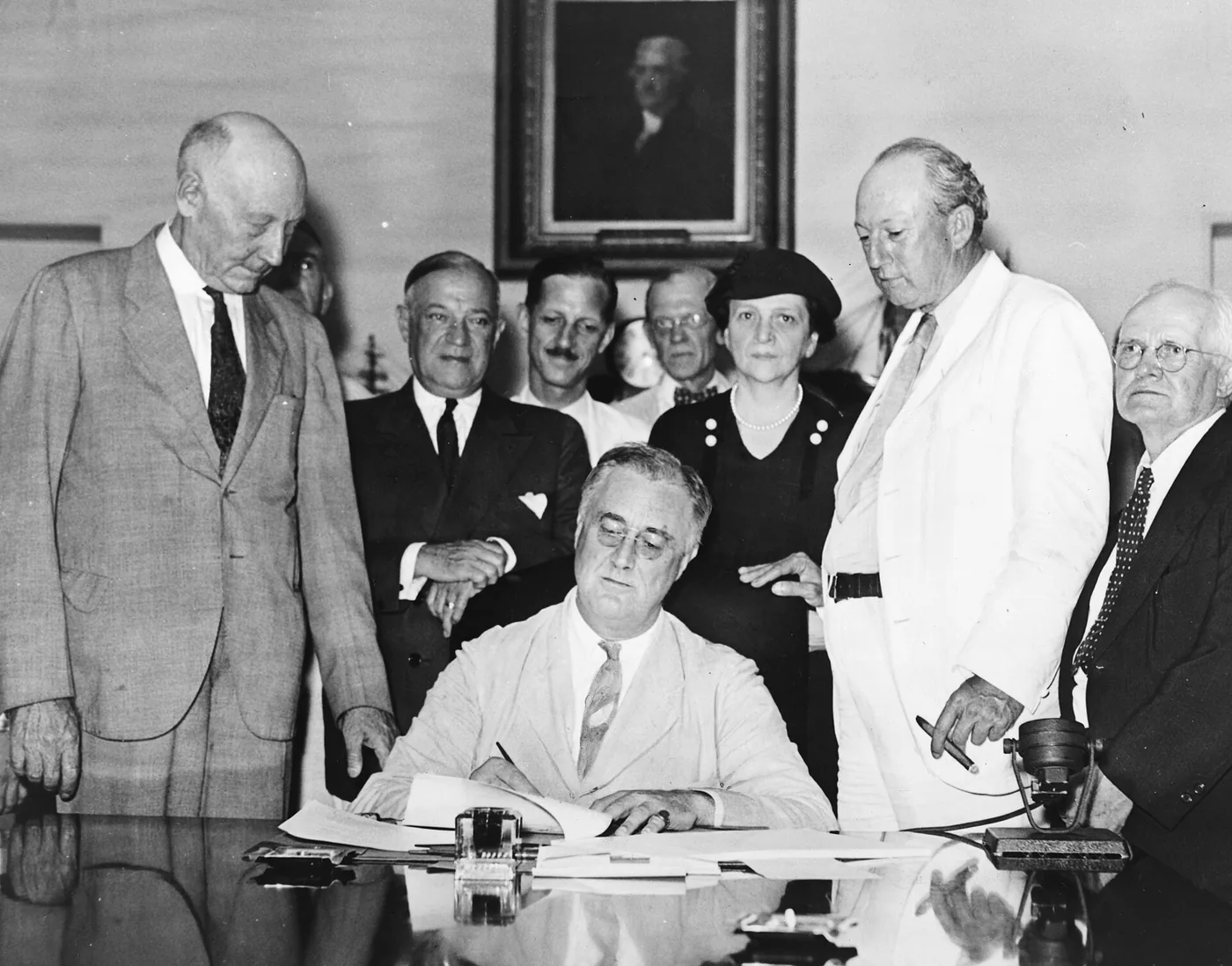 Black and white photograph of President Franklin Roosevelt seated at a desk signing the Social Security Act, surrounded by members of Congress and officials in suits.