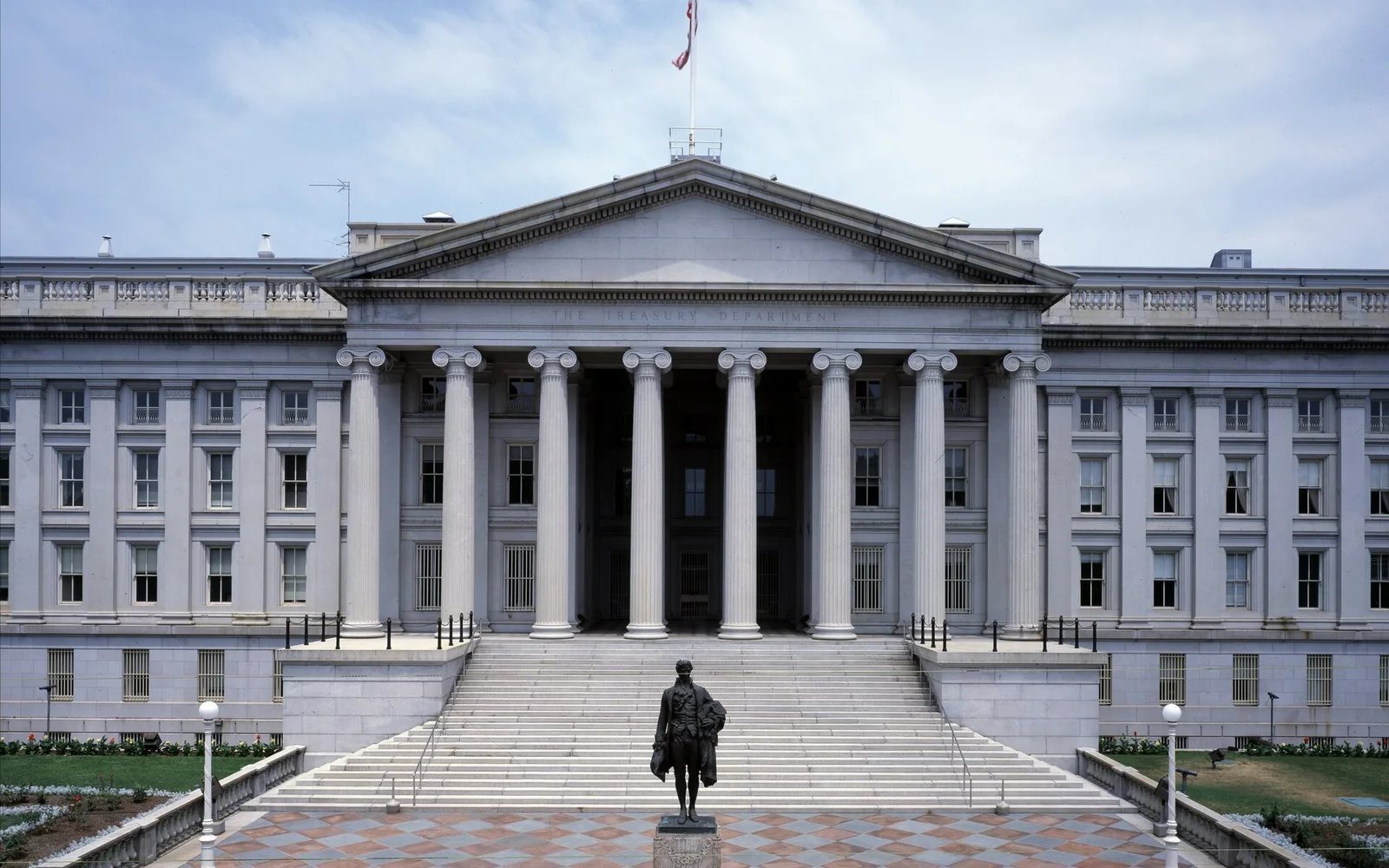 The columned south entrance of the U.S. Treasury Building in Washington, D.C., with a bronze statue of Alexander Hamilton on a plinth in the plaza in front and a flag on the roof.
